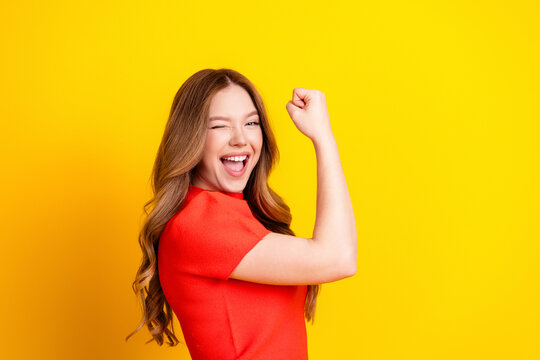 Smiling young woman in red celebrating victory and confidence against bright yellow background, winking and raising fist in joyful empowerment and success pose