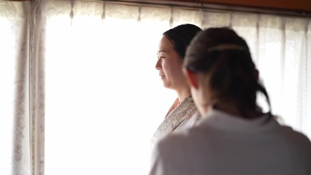 Soft light filters through lace curtains behind two women. One adjusts the other's kimono with gentle care. The second woman stands still, gazing forward