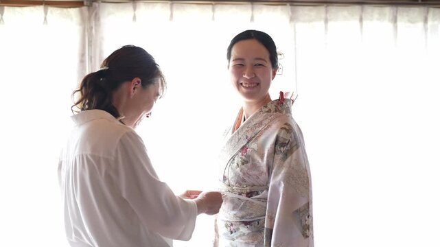 Soft light streams through sheer curtains. Woman adjusts a kimono's sash with gentle care. Another woman stands nearby, smiling warmly. Third person observes quietly, head bowed. Scene captures quiet