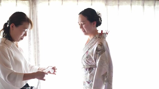 Soft light falls through curtains. Two women in white work quietly. One adjusts a kimono's sash. The other stands still, serene. Gentle hands and calm focus. Perfect for wedding or cultural scenes
