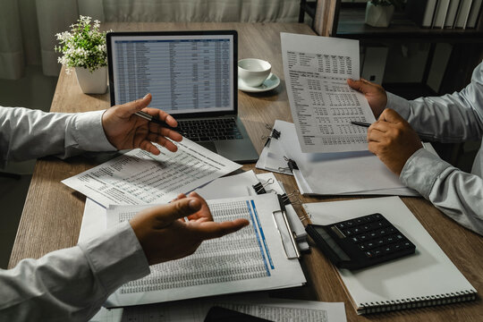 Coworkers discuss printed financial reports beside a laptop and calculator, brainstorming over spreadsheet figures and reviewing account records for collaborative business analysis.