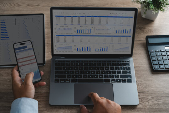 Hands use a laptop and smartphone beside a tablet and calculator, reviewing spreadsheet dashboards with charts for financial data analysis, revenue tracking, and business reporting.