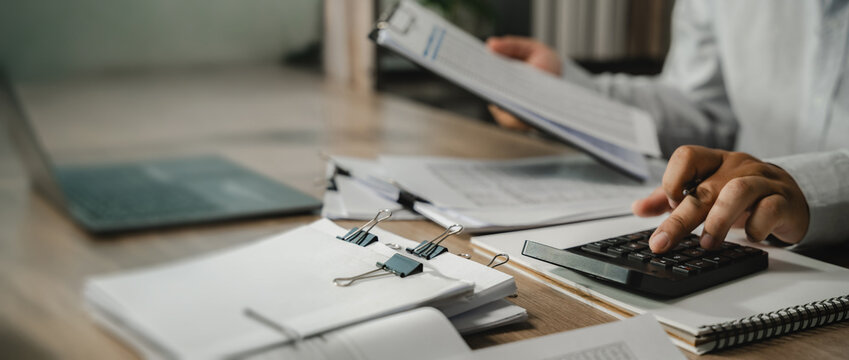 Accountant calculator while reviewing clipped financial papers near a laptop, checking budget figures and account records for finance analysis and business budgeting with copy space.