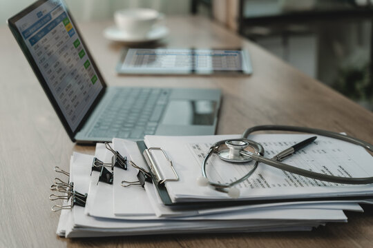 Stethoscope on clipboard with clipped paper documents beside laptop and tablet on wooden desk, showing medical records review, healthcare documentation, office workflow with copy space.