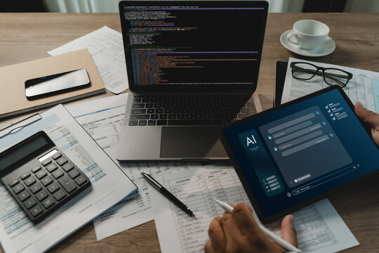 Businessman using a tablet with an AI chatbot interface beside a laptop with code, calculator and financial papers on a busy desk for digital document review and business analysis workflow.