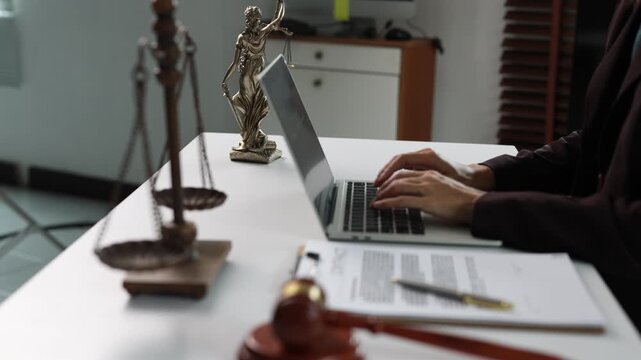 Female lawyer typing on a laptop in a law firm with a gavel, statue of Themis, and scales of justice on the desk while providing legal advice and online consultation