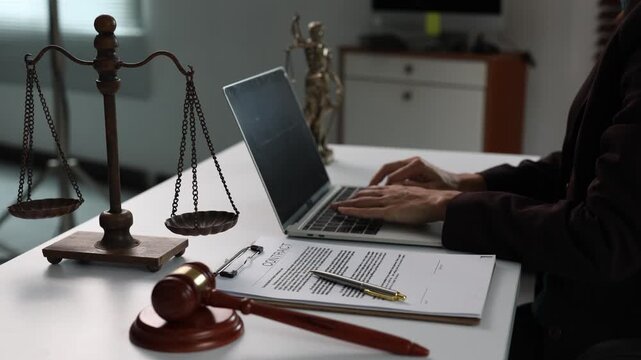 Professional lawyer working on a laptop in a courtroom or law firm office with a gavel, contract, scale of justice, and lady justice statue on the desk