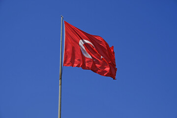 Turkish national flag on blue sky background. The flag on a flagpole flutters in the wind. The white star and crescent moon on red background. National identity background. © Yuliya