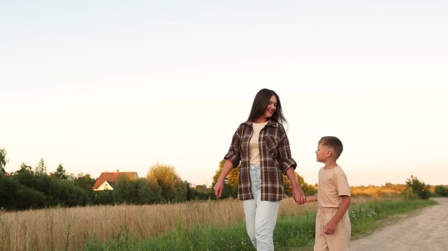 Happy mother and child walk hand in hand along sandy village path before sunset. Slow motion captures smiling faces and warm connection during peaceful evening.