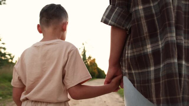 Close up shows mother holding child hand while walking along sandy path toward horizon. Slow motion captures gentle movement and peaceful evening in nature at sunset.