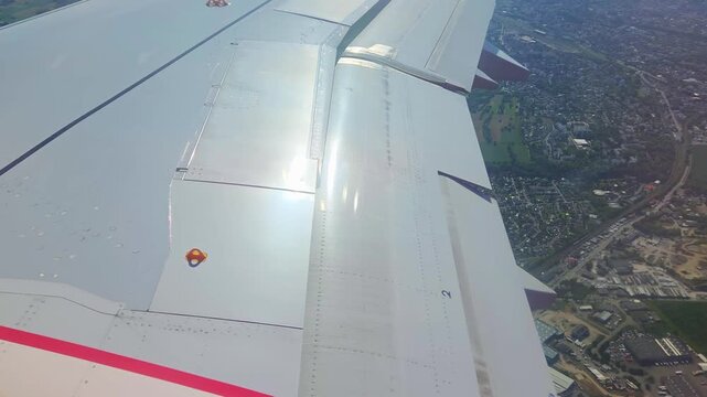 Aircraft wing with visible flaps and ailerons in motion during descent, showcasing landscape below with fields and buildings as the plane approaches landing