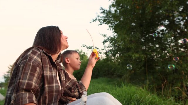 Happy mother and son sit on grass blowing soap bubbles at sunset. Slow motion shows family connection peaceful childhood and warm outdoor lifestyle.