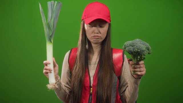 Young chinese woman courier wearing red cap and backpack holds a leek and a broccoli, pouting with closed eyes in a studio; food choice frustration.