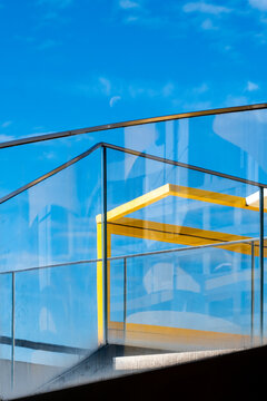 Against vivid blue sky, transparent planes catch the light as modern architecture reveals a glass staircase, its yellow railing forming a geometric reflection