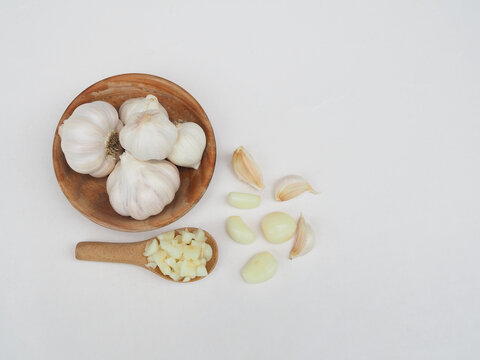 Chopped garlic is in a wooden spoon and garlic is in a wooden cup, placed on a white surface.