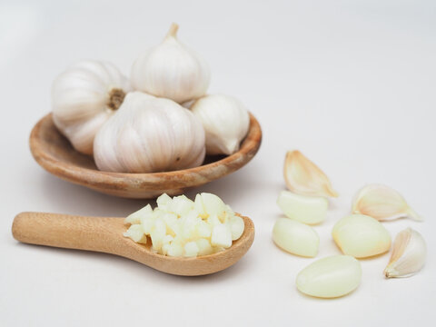 Chopped garlic is in a wooden spoon and garlic is in a wooden cup, placed on a white surface.