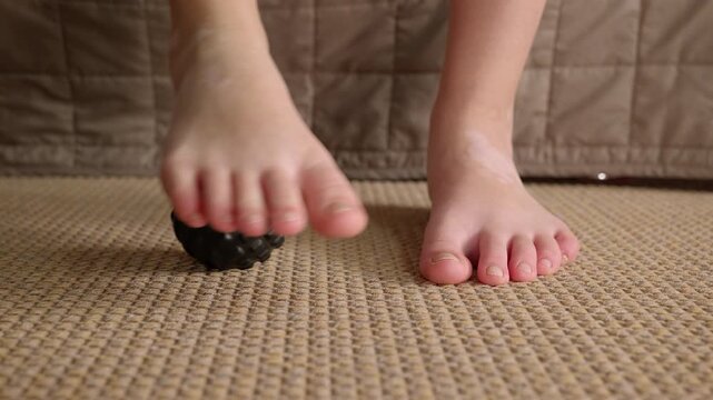 Person massaging feet with spiky ball for plantar fasciitis. Bare feet using a black spiky massage ball on a carpet to relieve foot pain and treat plantar fasciitis
