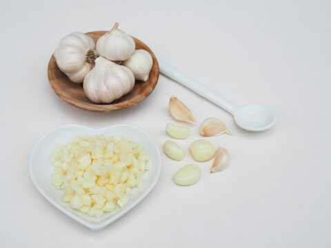 Chopped garlic is in a wooden spoon and garlic is in a wooden cup, placed on a white surface.
