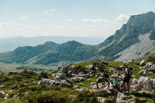Female cyclist standing beside a gravel bike overlooking Sedlo Pass in Durmitor National Park, Montenegro. Rocky alpine landscape and towering peaks dominate the panoramic view