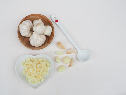 Chopped garlic is in a wooden spoon and garlic is in a wooden cup, placed on a white surface.