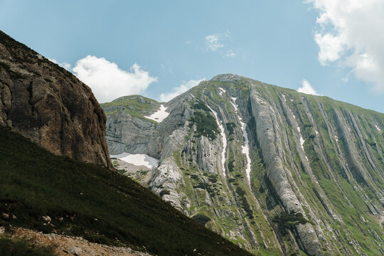 Towering limestone cliffs with lingering snowfields dominate the landscape in Durmitor National Park, Montenegro. Rugged alpine scenery rises beneath a bright blue summer sky