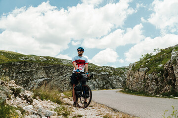 Fototapeta premium Bikepacking cyclist pauses beside a winding mountain road in Durmitor National Park, Montenegro. Loaded gravel bike and rugged alpine scenery capture the spirit of adventure travel