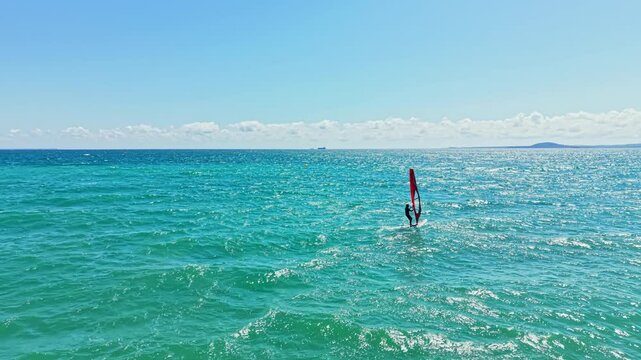 A person is windsurfing in the ocean. Waves move smoothly under the board as the wind carries the sail. The sun shines brightly and the sky is mostly clear with few clouds.