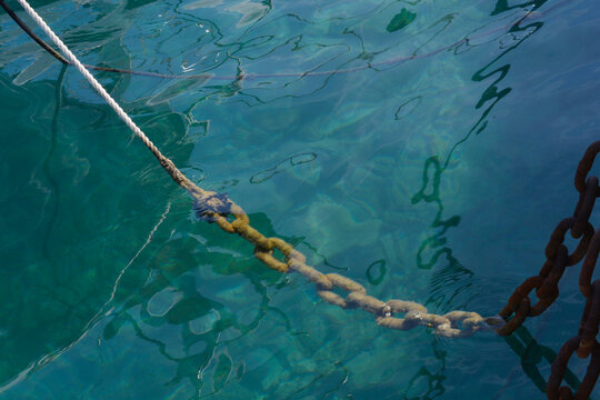A thick rusty anchor chain connected to a white mooring rope descends into clear blue sea water with surface ripples and reflections