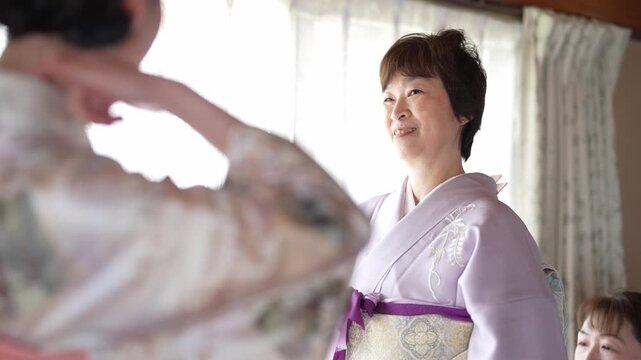 Soft light illuminates a serene dressing room. Two women share a joyful moment in traditional attire. One laughs, her eyes sparkling with delight. The other smiles warmly, adjusting a purple sash