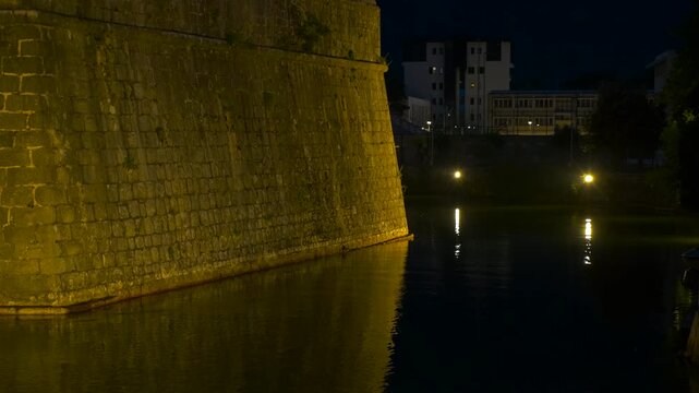 Medieval castle moat with golden light reflections at night. Ancient stone fortress walls illuminated by warm golden lights, creating mesmerizing reflections on the dark, tranquil water