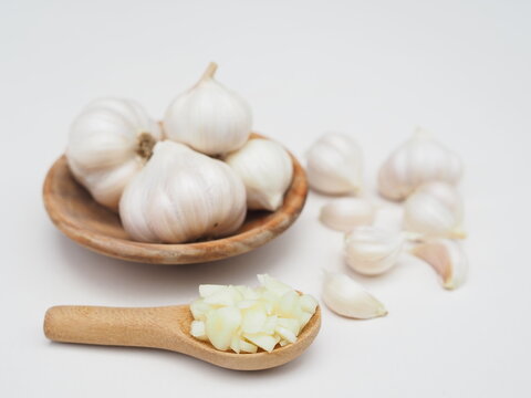 Chopped garlic is in a wooden spoon and garlic is in a wooden cup, placed on a white surface.