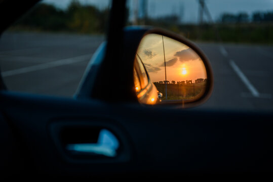 Right side rearview mirror of a car reflecting sunset. Close-up view of the vehicle's exterior right mirror with sunset sky and landscape reflection.