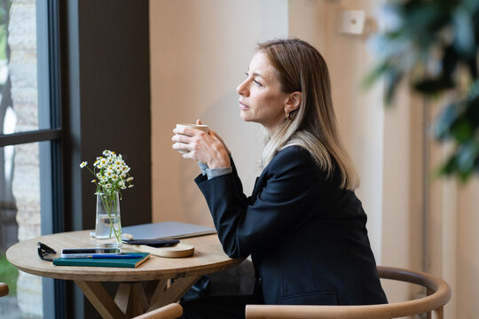Pensive businesswoman takes coffee break in cafe, looks out window. Sad woman experiencing emotional burnout from multitasking at work, sitting in cafeteria alone, thinking about life troubles. 
