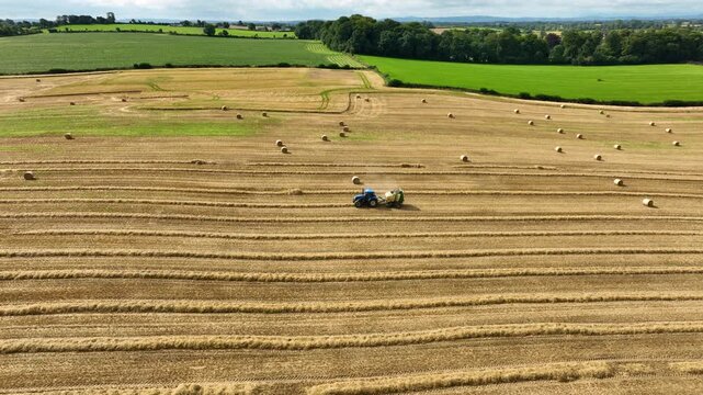Tractor Pulling Round Baler Across Wide Countryside Field Dotted With Hay Bales
