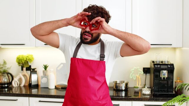 Arabian man jokes with pepper rings on eyes in kitchen while preparing salad for healthy keto eating. Young guy laughs feeling playful and satisfied for real clean nutrition at home apartment indoors.