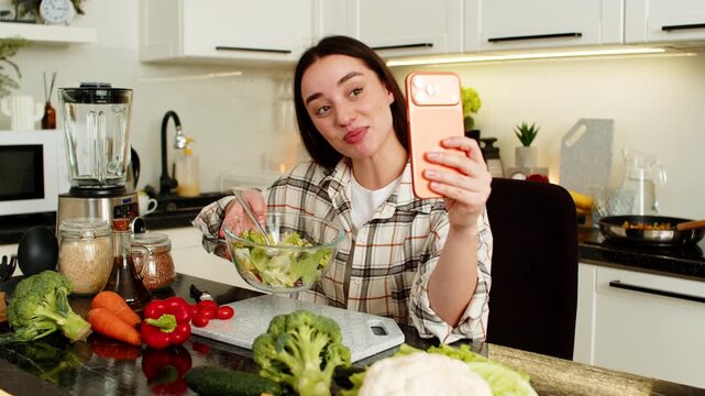 Young woman blogger holds orange smartphone and records keto diet vlog while mixing veggie salad. Healthy cooking content for social media, home kitchen lifestyle, fresh low carb ingredients on table.