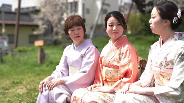 Three women in traditional Japanese kimonos sit together outdoors. They wear soft pastel colors and floral patterns. Their expressions show quiet happiness and contentment