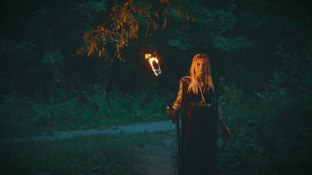 Gothic fantasy woman dark queen walking in mystical night forest trees, holding burning torch in hands, bright fire flame lights way. Sexy fairy Girl shaman black long boho style dress beauty face