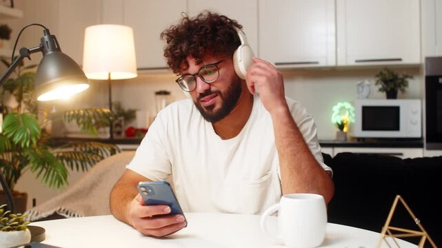 Arabian man at home wears wireless headphones listens music nods slowly enjoys calm, dancing. Egyptian guy sitting at table rests near laptop switches tracks breath steady quiet calm relax tonight.