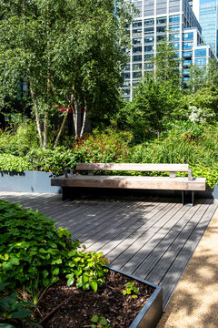 A long bench anchors a garden terrace in shade, where trees and greenery surround layered plants as the city landscape meets contemporary architecture