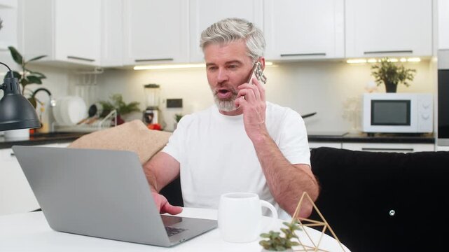 Middle-aged man at home sitting at table works on laptop and talks on smartphone to client. Mature guy on sofa types replies speaks steady voice manages task list and keeps focus under load clear tone