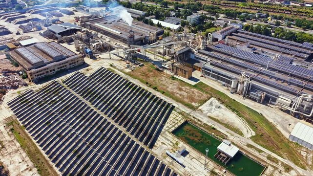 Solar panels are placed in rows on the ground near industrial buildings. Smoke rises from chimneys while the sun is shining. This scene shows a mix of energy sources and industry.