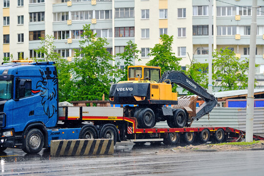 Minsk, Belarus. May 13, 2026. Scania truck with flatbed trailer transporting yellow black Volvo excavator on wet urban street to construction site with apartment buildings. Heavy equipment haulage