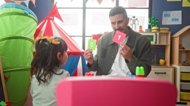 Male teacher holds number flashcards and points with hand while a young student watches across a play table in a playroom building; focused learning.