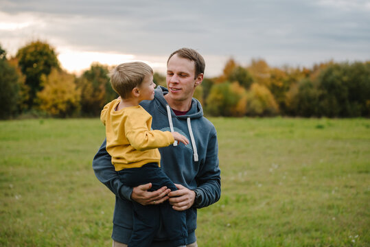 Photo portrait of dad holding little boy outdoors in autumn field. Father and son looking at each other and smiling during a walk in nature.