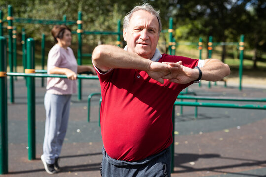 Staying after retirement. Happy joyful mature retired sportsman wearing sportswear doing side stretching exercises with arm over his head, exercising outside in city park