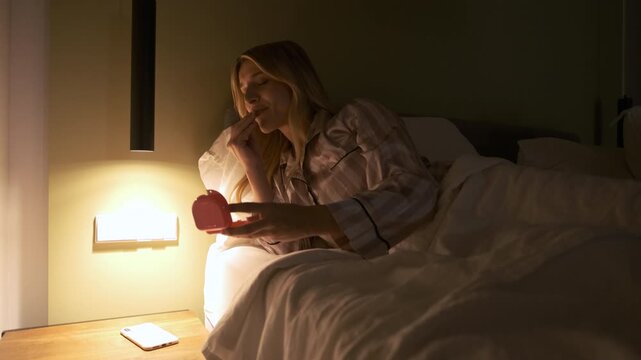 Young transgender woman putting on transparent teeth aligners while reclining on bed before going to sleep in dark bedroom