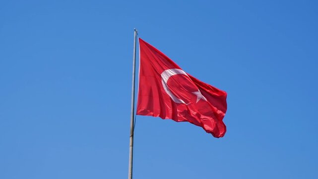 Turkish national flag on blue sky background. The flag on a flagpole flutters in the wind. The white star and crescent moon on red background. National identity background.