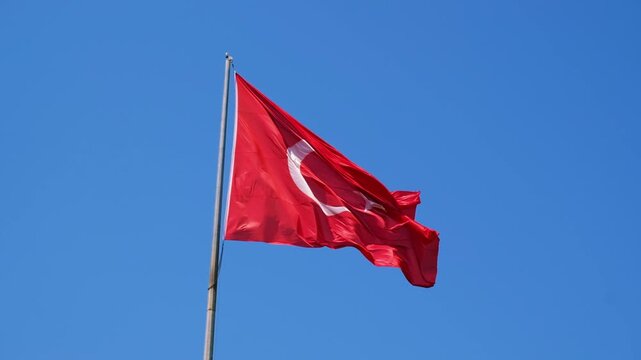 Turkish national flag on blue sky background. The flag on a flagpole flutters in the wind. The white star and crescent moon on red background. National identity background.