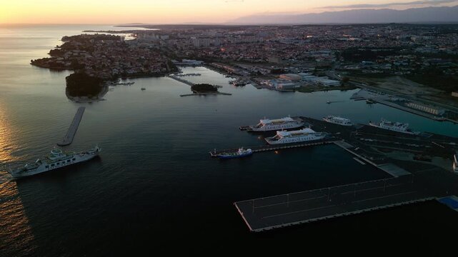 Aerial footage of Hel Peninsula harbor in Poland at sunset, ferries docked along calm reflective waters, golden evening sky, and the town stretching across the narrow coastline.